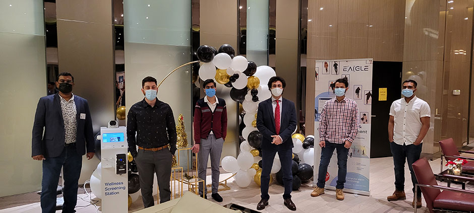 Six EAIGLE staff members at their Ontario headquarters, wearing masks and standing socially distanced from each other, next to a Wellness Screening Station