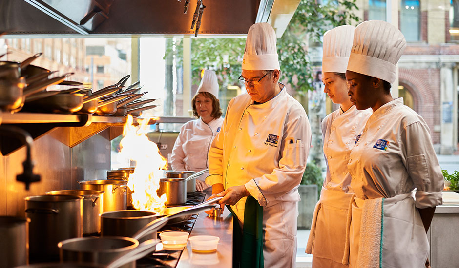 Des étudiants en cuisine observent leur professeur en train de manipuler un plat enflammé.