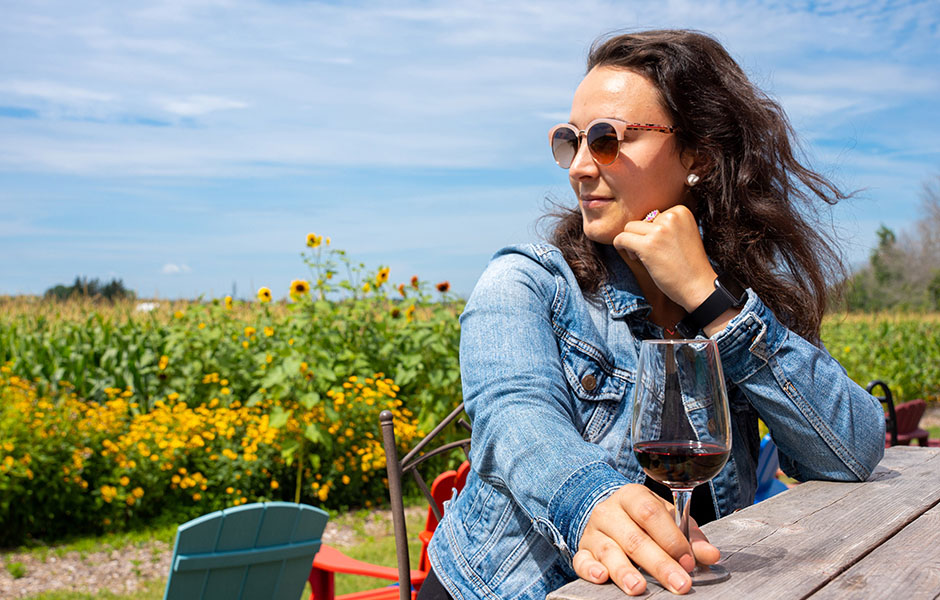 Woman sitting at a picnic bench with a glass of red wine, gazing at a field of sunflowers.